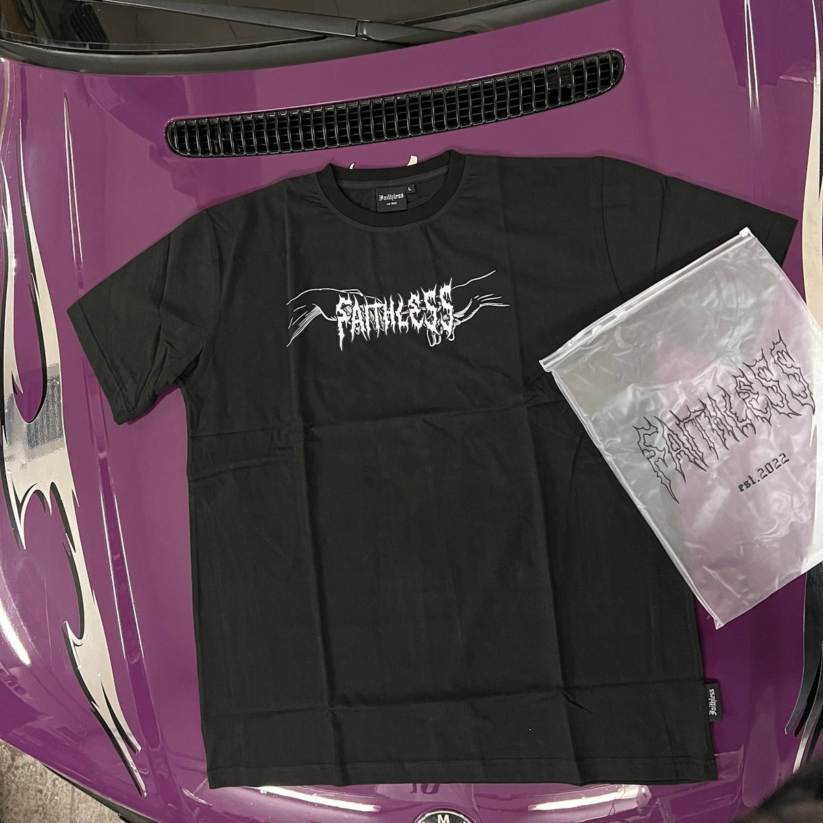 Black Angel tee, front view with white Faithless chest print, on a purple car hood next to its Faithless bag